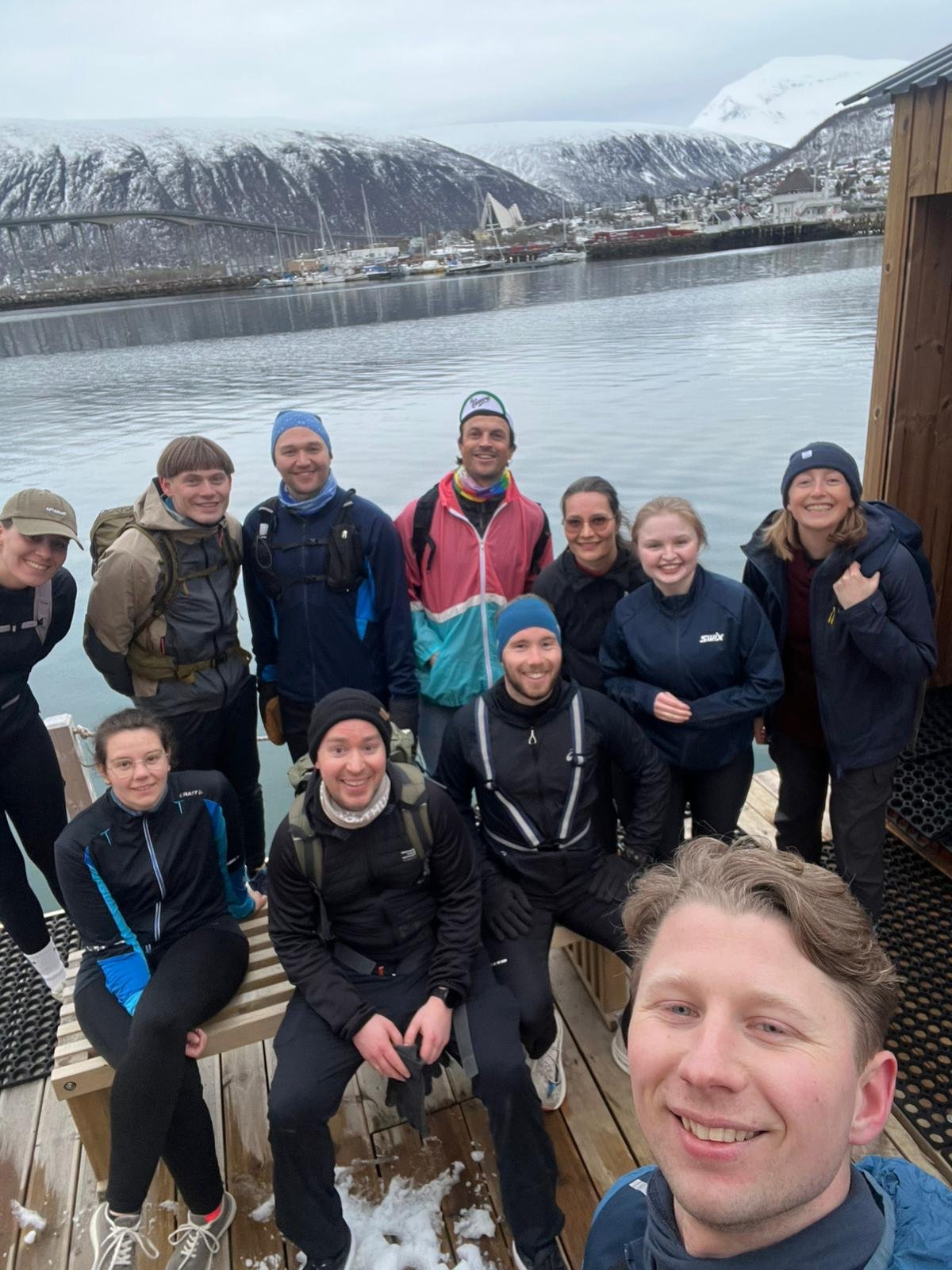 A smiling group of guests and guides gathered on a jetty in Tromsø, with snow-covered mountains, the fjord and the Arctic Cathedral behind them