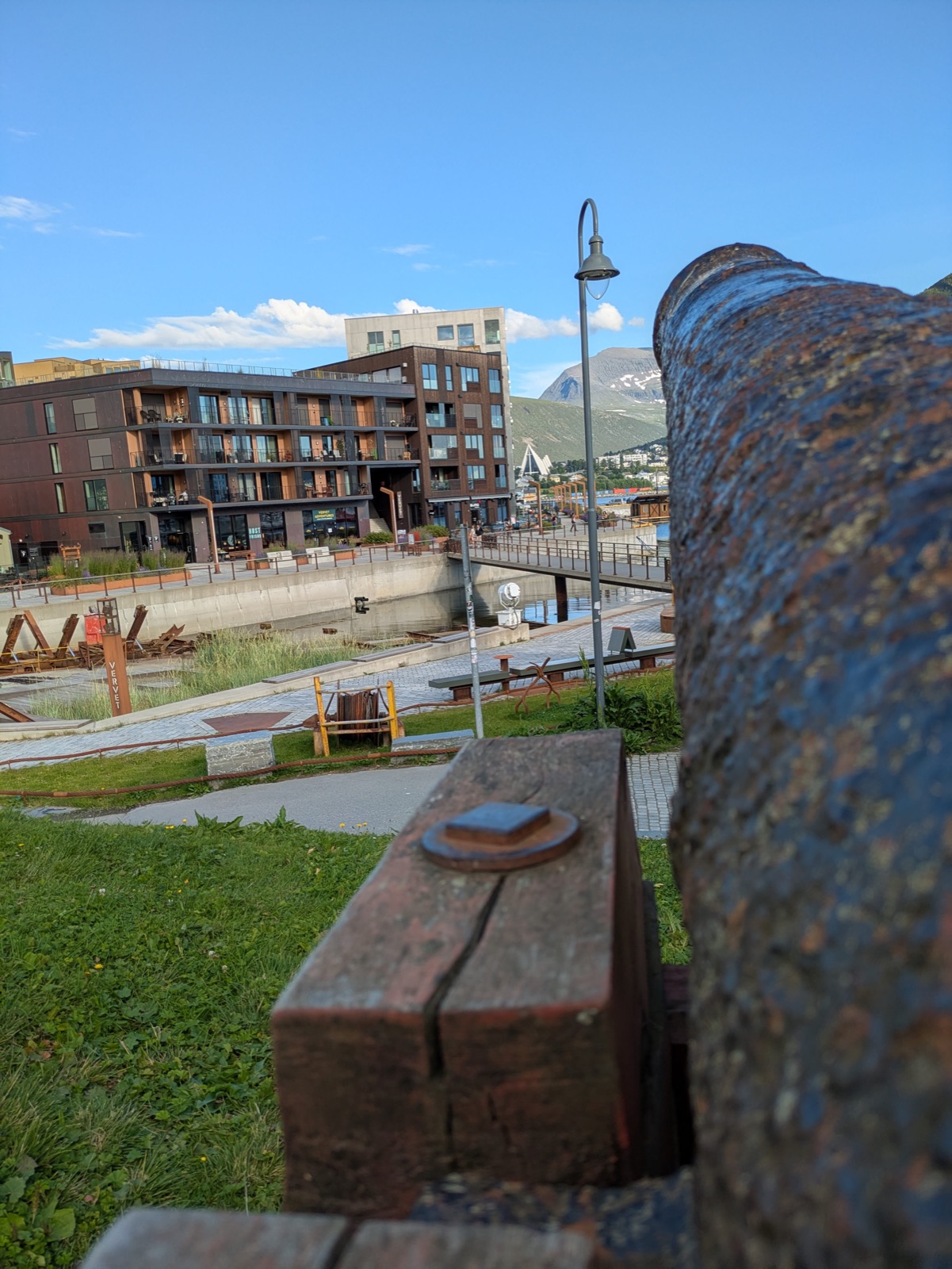 A rusty old cannon on the Tromsø waterfront with modern harbour buildings, a mountain and the Arctic Cathedral in the distance